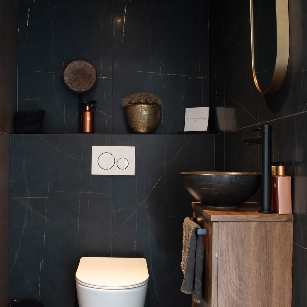 Toilet met donkere marmerlookwanden, fonteinkastje in houttinten, zwarte waskom en spiegel met goudkleurige rand.