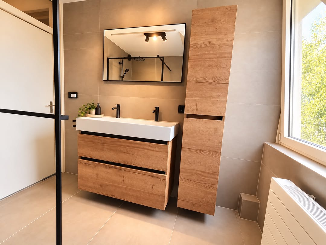 Badkamer met beige tegels, zwevend houtlook wastafelmeubel, hoge kast, zwarte kranen en een zwart omlijste spiegel.