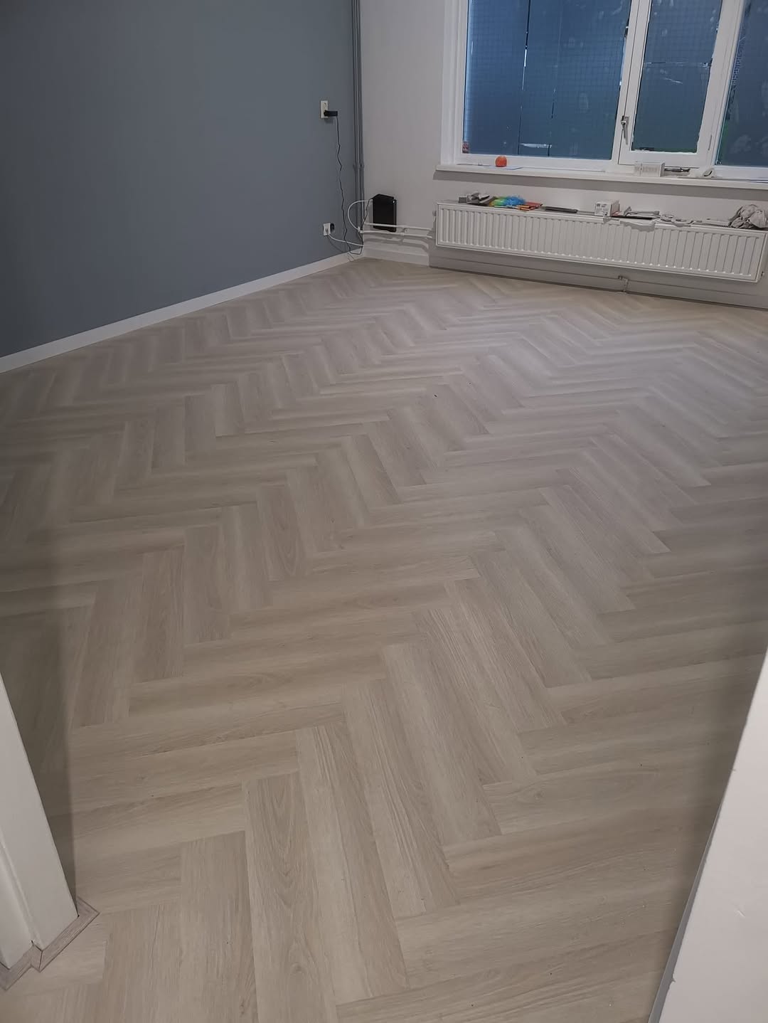 Lichte PVC-clickvisgraatvloer met houtlook in een ruimte met een blauwgrijze wand en witte plinten.