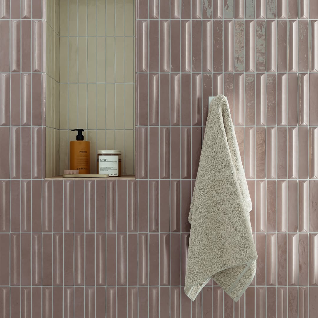 Zachtroze wandtegels met reliëf, een licht betegelde nis met verzorgingsproducten en een beige handdoek.