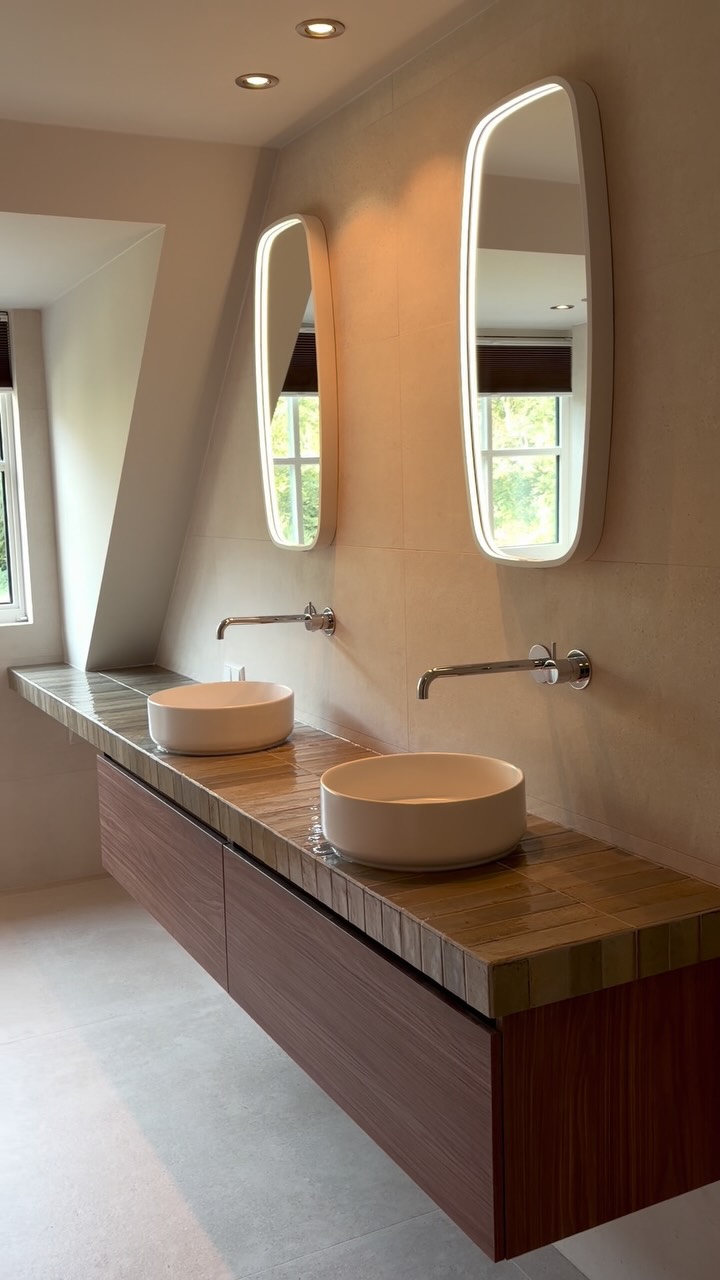 Badkamer met betegeld wastafelblad, twee ronde waskommen, meubel met houtlook en langwerpige verlichte spiegels.
