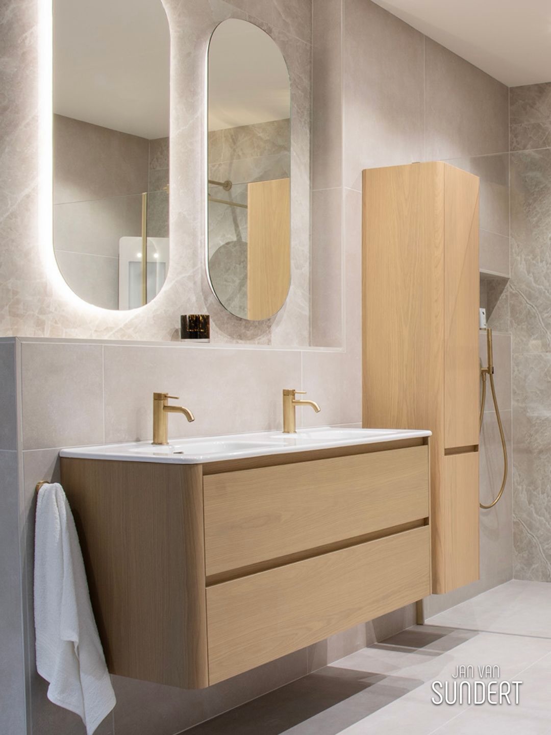 Badkamer met licht houten wastafelmeubel, zandkleurige tegels, afgeronde spiegels en champagnekleurige kranen.