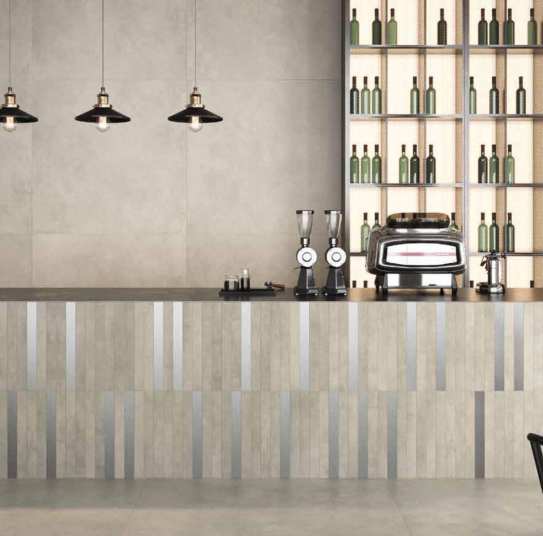 Barinterieur met grijze betonlooktegels, een front met verticale stroken, zwarte hanglampen en verlichte flessenrekken.